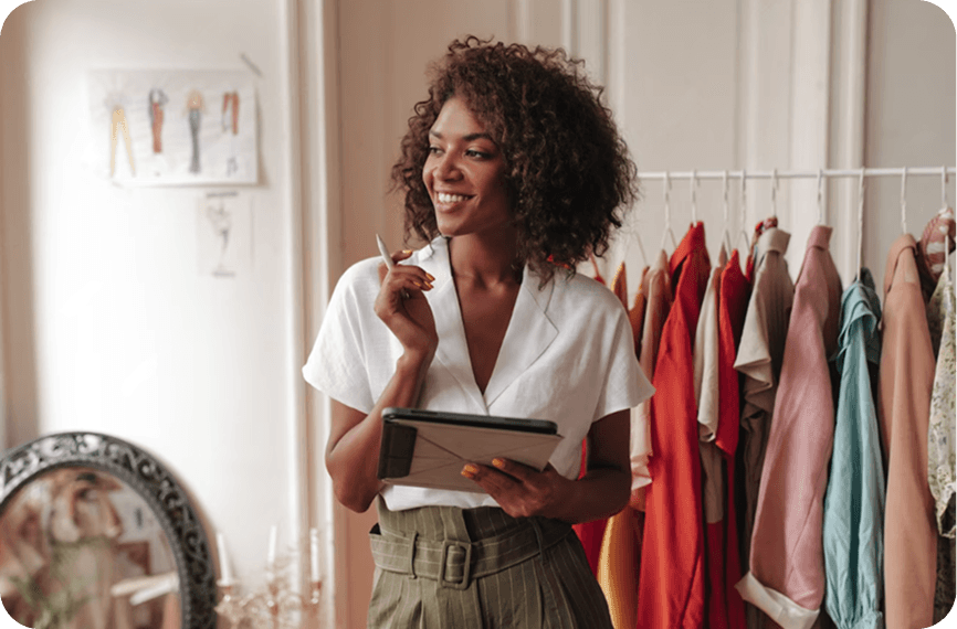 Fashion Designer Working on Fashion Collection In Silver Spoon A designer with a thoughtful expression jots down notes using Silver Spoon, while surrounded by clothing racks in a bright studio.